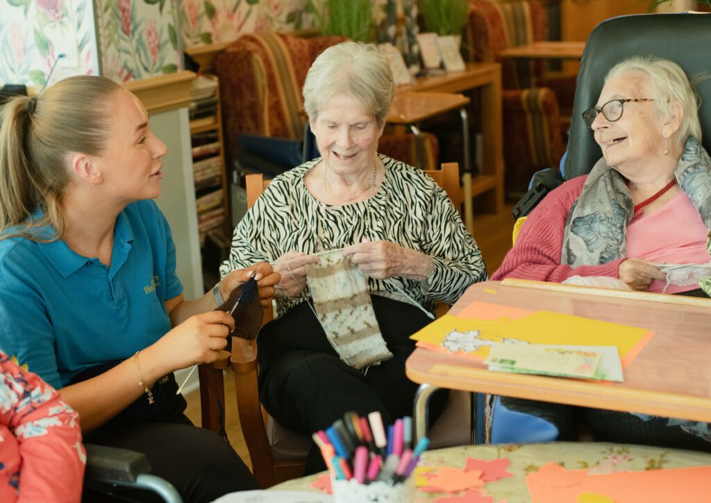 Two seniors knitting and sitting with a resident caregiver.